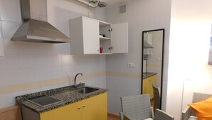 A compact kitchen with a granite countertop, a stainless steel sink, and a built-in oven. There is a wicker chair and a small table with an orange cushion. - Cádiz INN Hostel (Cádiz)