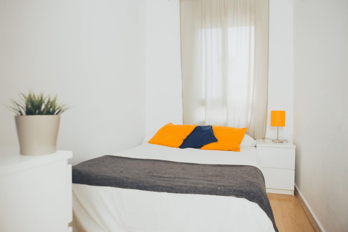 Quarto com cama, mesa de cabeceira com abajur, planta em vaso e janela com cortinas leves.