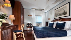 A hotel room with a large bed, a desk with a lamp, a chair, a small table with a suitcase, and a view of the sea. - Hotel Atilius (Riccione)