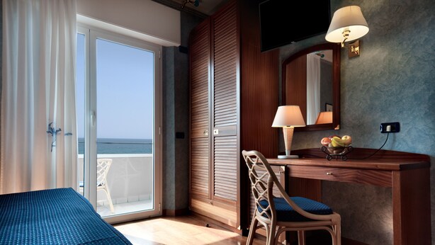 A room with a bed, a desk with a chair, a television, and a view of the sea. - Hotel Atilius (Riccione)
