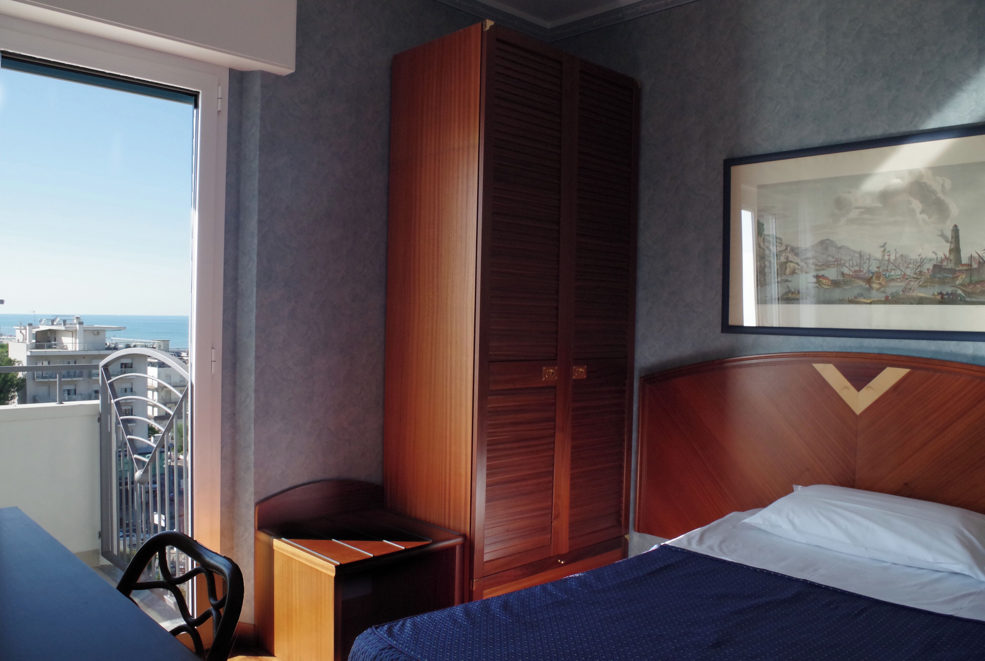 Une chambre d’hôtel avec un lit, une armoire en bois, un tableau encadré au mur et une vue sur la ville par la fenêtre.
