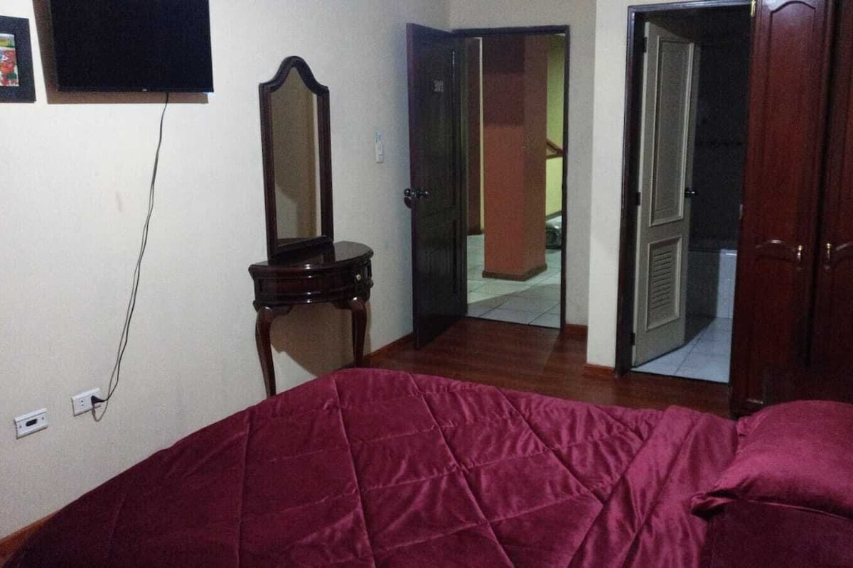 Quarto com cama, penteadeira, espelho e porta que dá para outro cômodo.