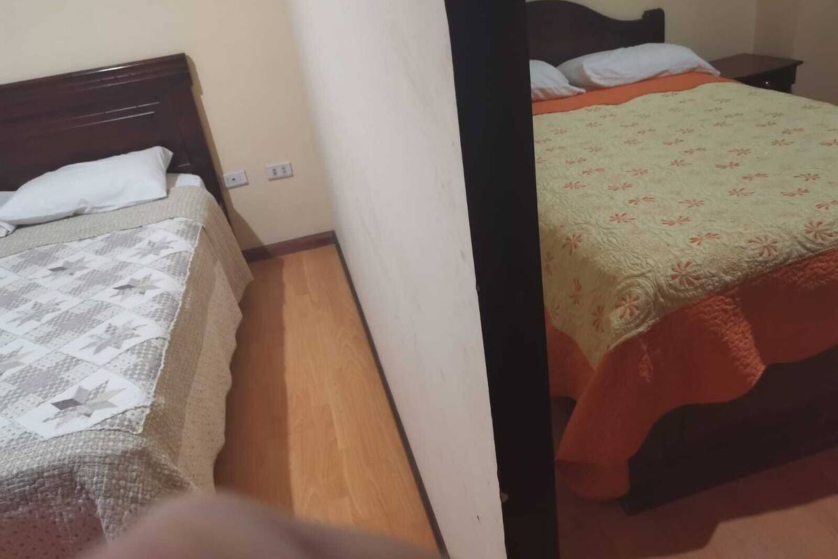 Quarto com duas camas, cada uma com estilo de roupa de cama diferente. O quarto possui piso de madeira e paredes brancas.