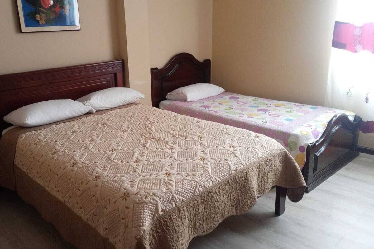 Quarto com duas camas, cada uma com cabeceira de madeira e colcha estampada. Há uma mesinha de cabeceira com gaveta do lado esquerdo.