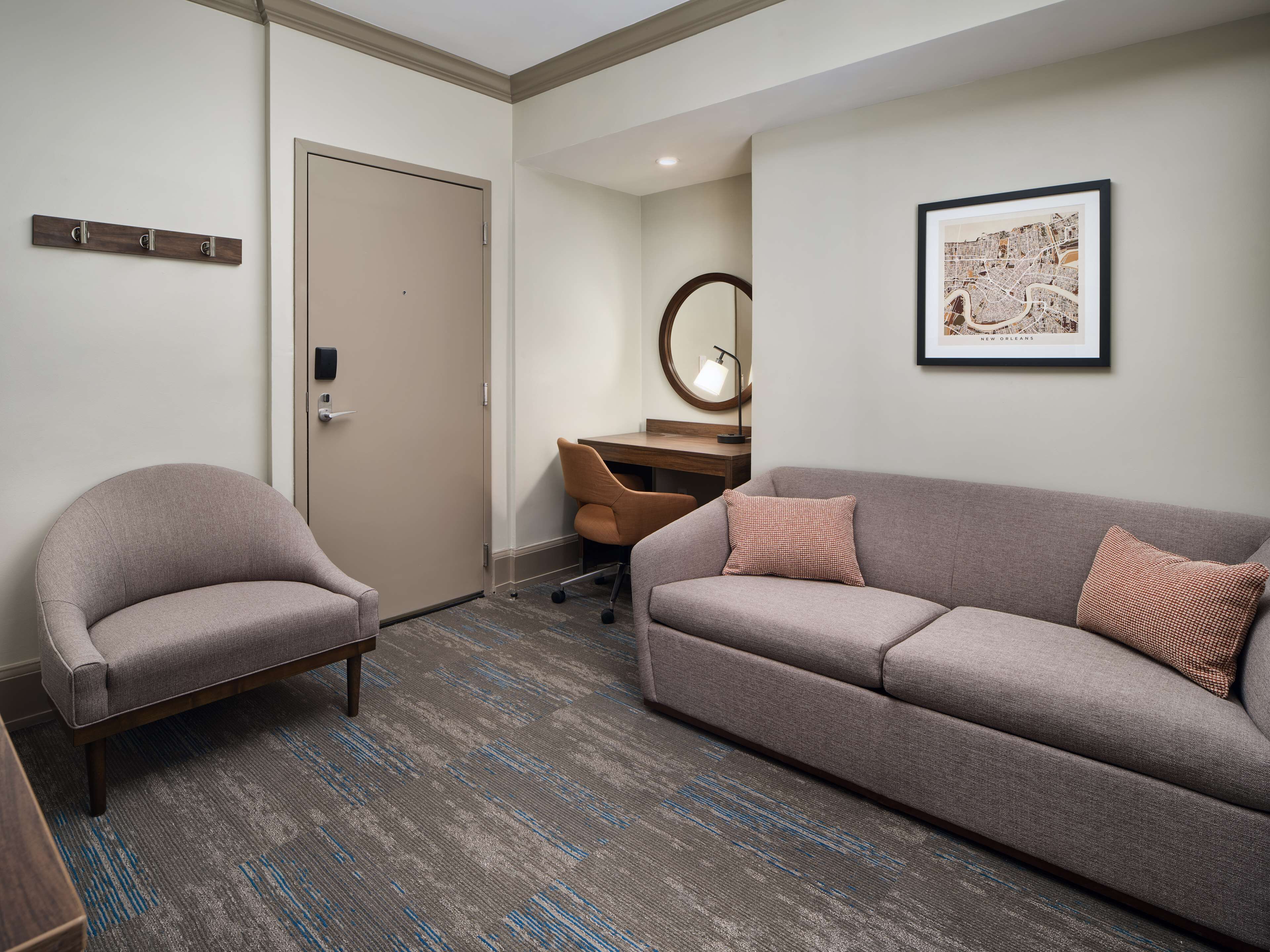 A hotel room with a grey sofa, a chair, a desk with a lamp, a framed map on the wall, and a door.
