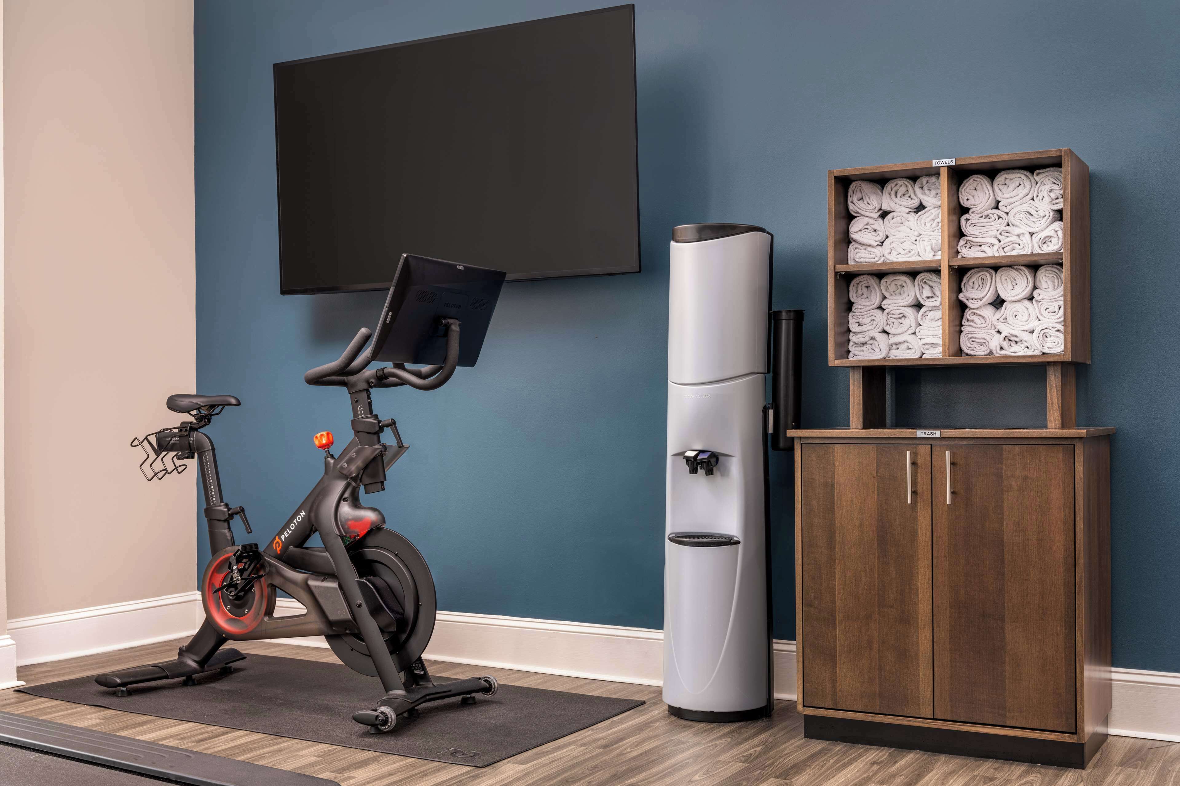 Una habitación con una bicicleta estática, dispensador de agua, un televisor y un armario con toallas dobladas.