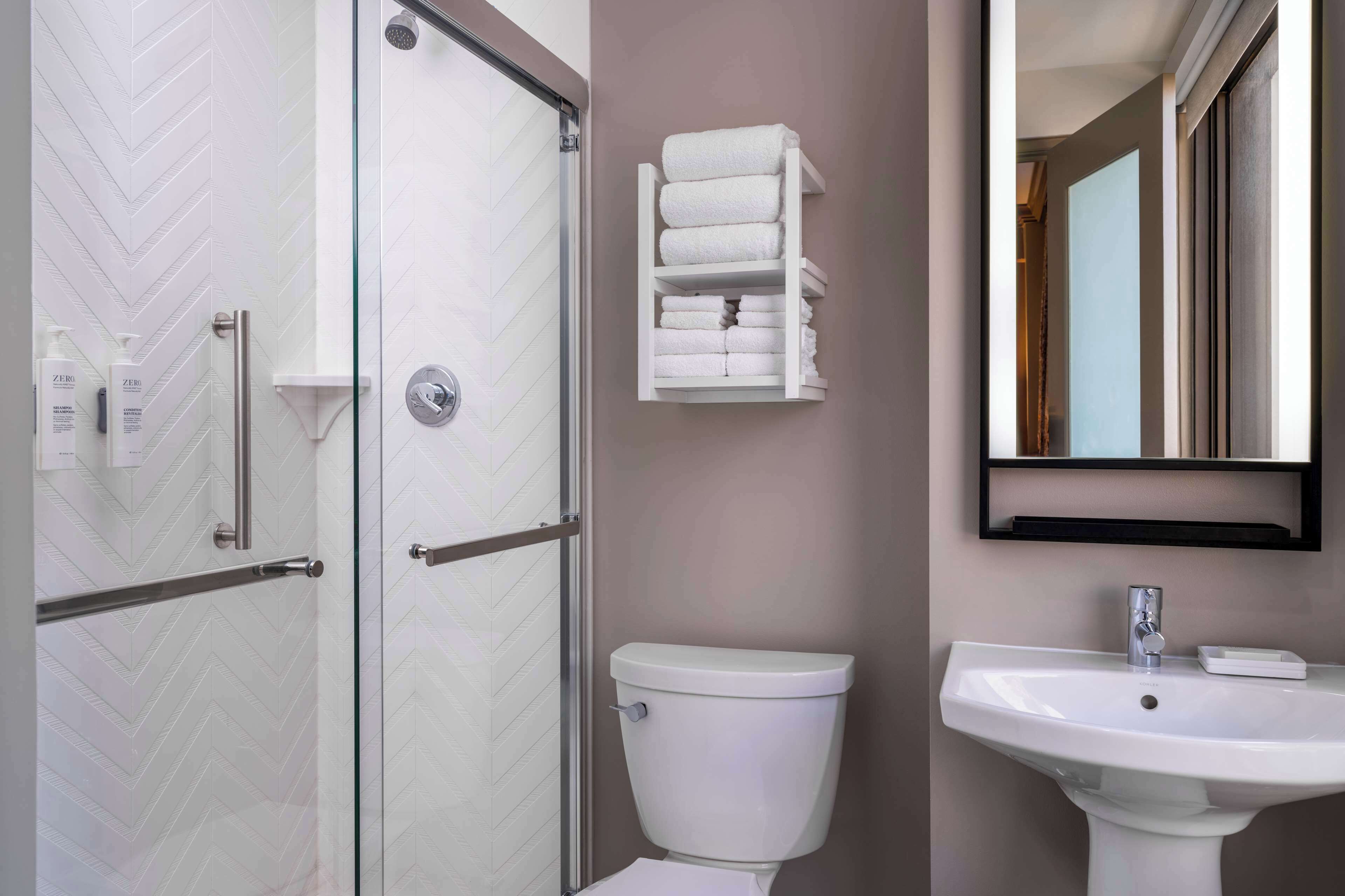 A bathroom with a glass shower enclosure, a white toilet, a sink with a mirror above it, and a towel rack with folded towels.