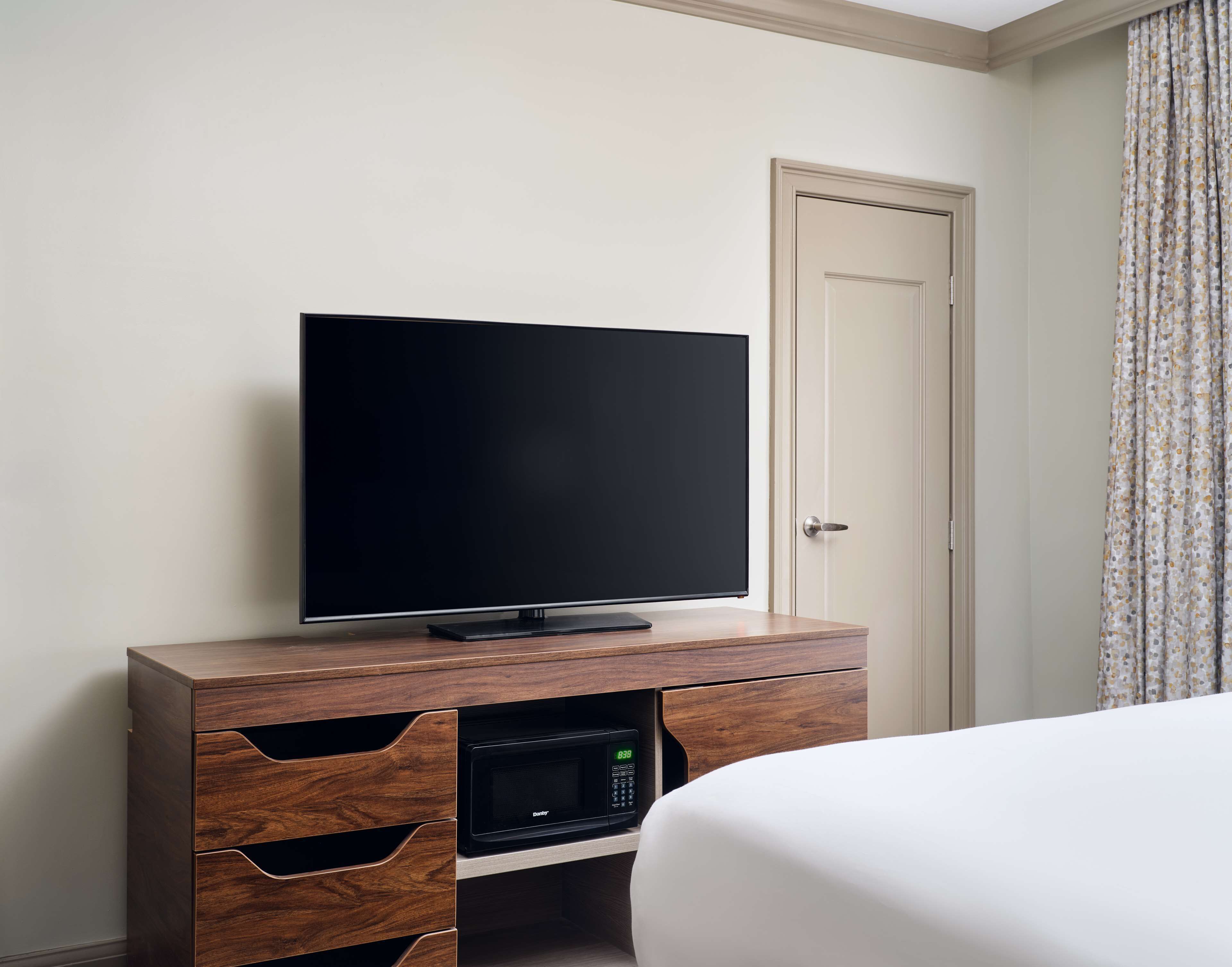 Une chambre d’hôtel équipée d’un téléviseur à écran plat posé sur un meuble en bois, d’un four à micro-ondes et d’une porte ornée d’un rideau à motifs.
