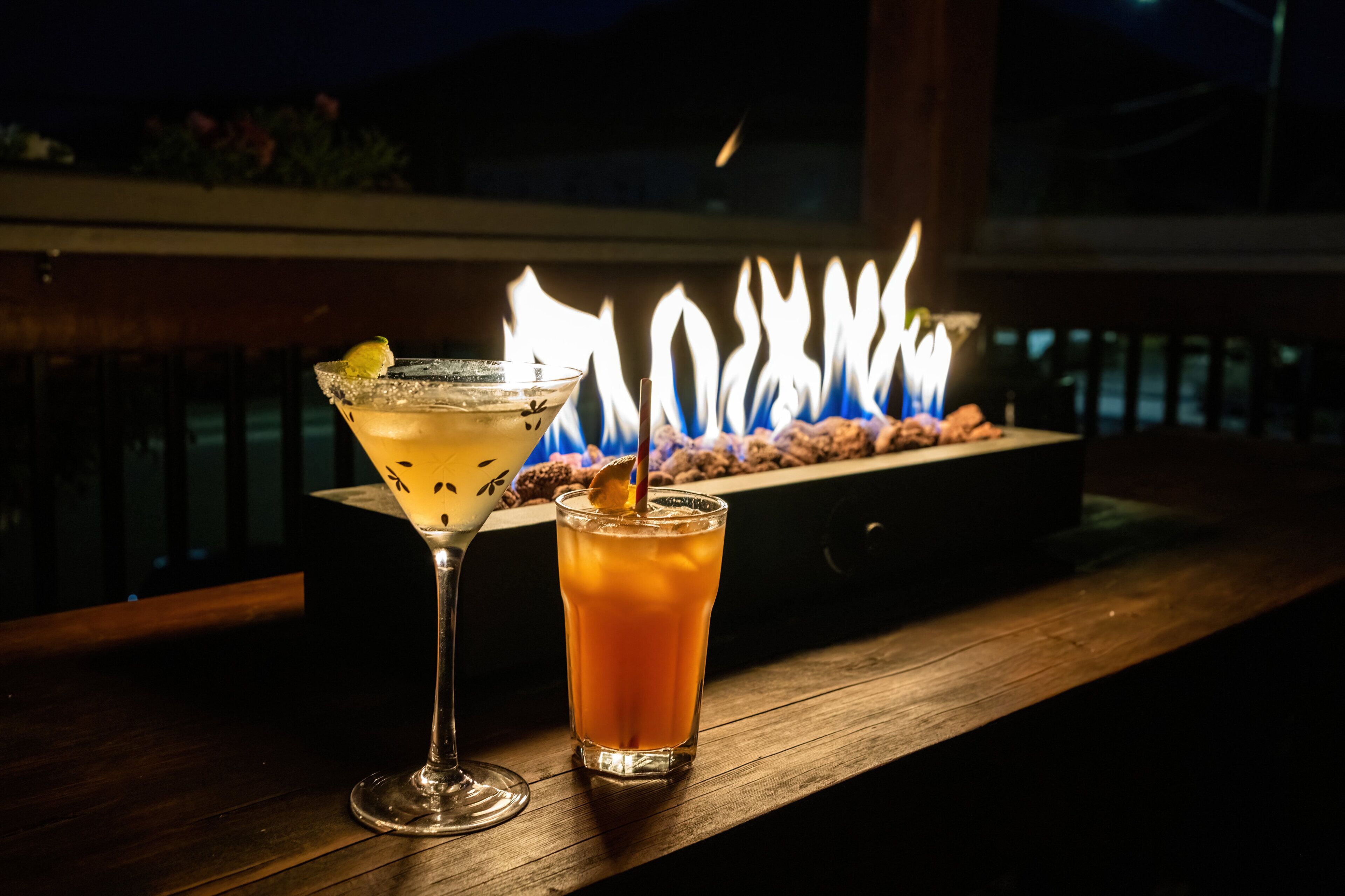 Two drinks on a wooden table with a fire in the background.