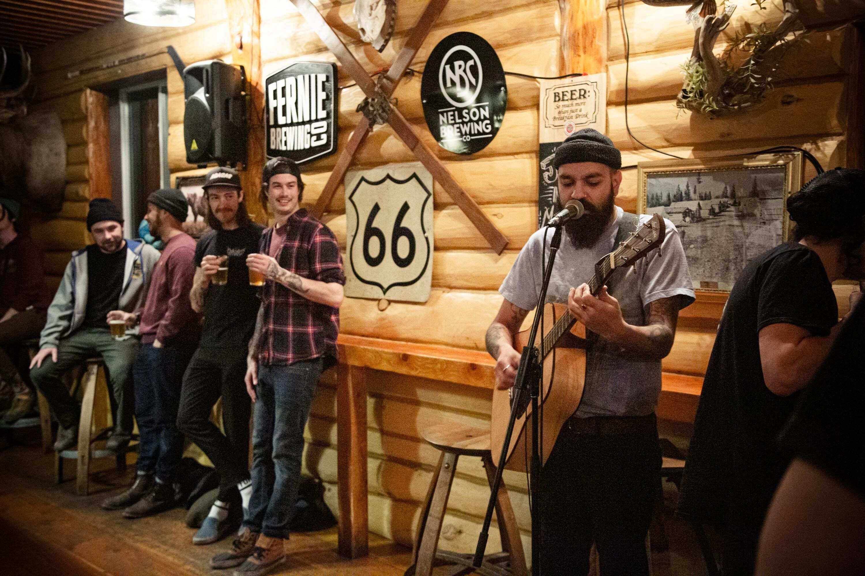 A rustic wooden interior with a musician performing, surrounded by patrons. Visible signs include "FERNIE BREWIES" and "NELSON BREWING."