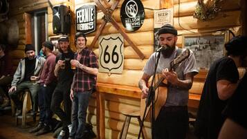 A rustic wooden interior with a musician performing, surrounded by patrons. Visible signs include "FERNIE BREWIES" and "NELSON BREWING."