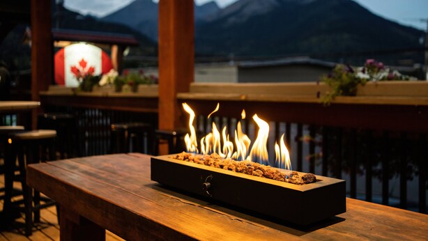 A wooden table with a built-in fire feature, set against a backdrop of mountains and a lit sign with a maple leaf.
