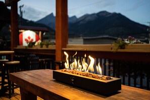A wooden table with a built-in fire feature, set against a backdrop of mountains and a lit sign with a maple leaf.