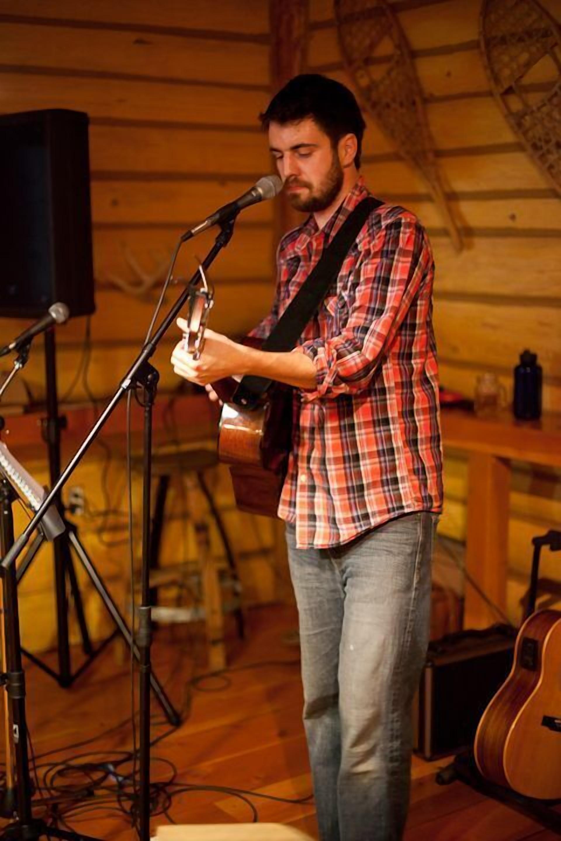 A person playing a guitar at a microphone stand in a room with wooden walls and a guitar in the background.