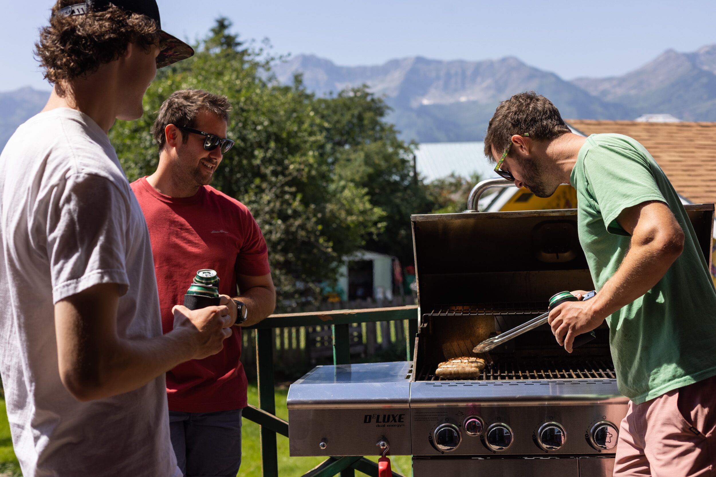 A group of people around a D'Luxe grill outdoors with a mountainous backdrop.