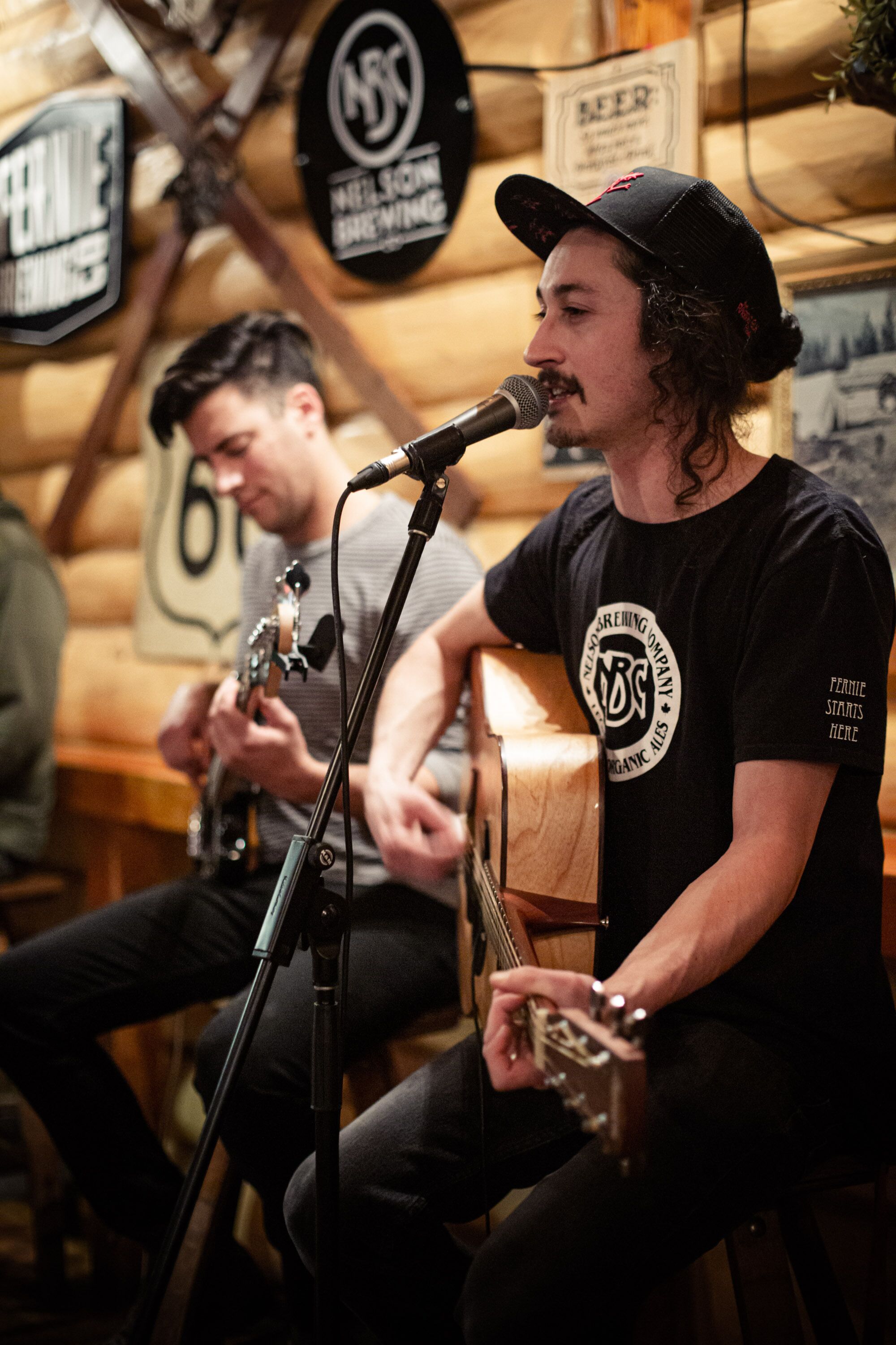 Un musicien joue de la guitare et chante dans un microphone, dans un cadre rustique aux murs en bois, avec une enseigne indiquant « Bière ».