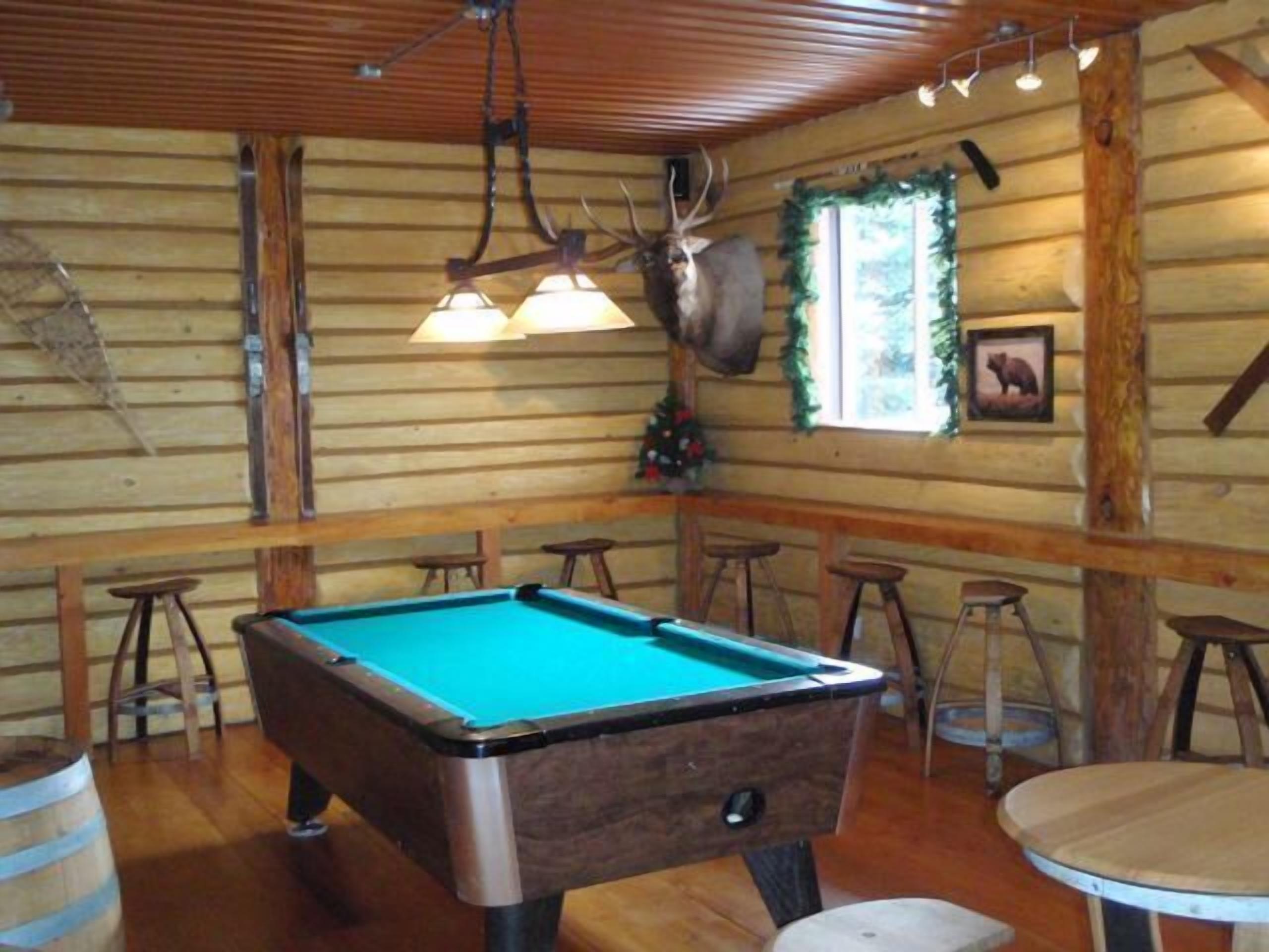 A pool table in a wooden cabin with a ceiling light fixture, wooden walls, and a mounted deer head.