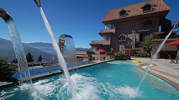A pool with multiple water jets, a clear blue sky, and a building with a red roof in the background. - Chun Jing Hillview Villa (Ren'ai)