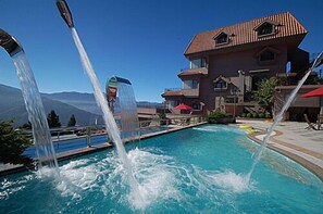Una piscina con múltiples chorros de agua, un cielo azul despejado y un edificio con techo rojo al fondo.