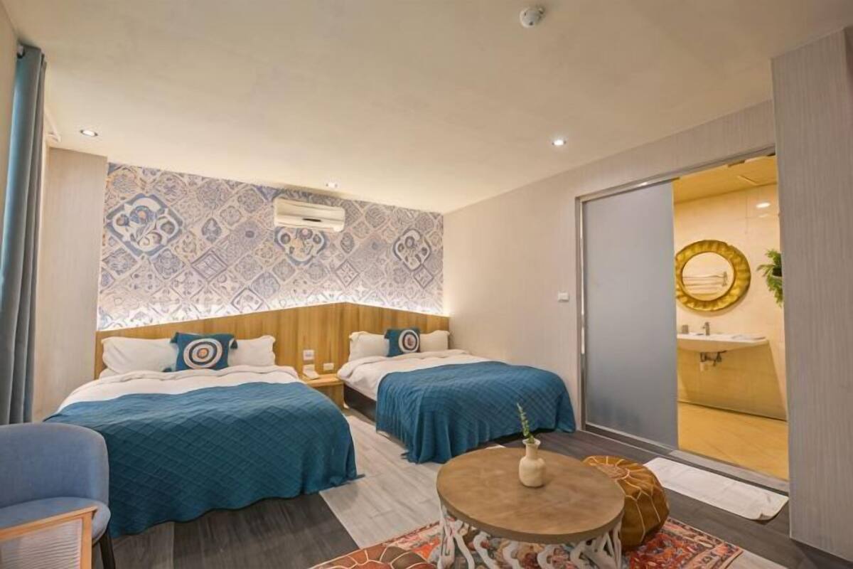 Ein Hotelzimmer mit zwei Betten, einem runden Tisch, einer Couch, einer Wand mit Muster und einem durch eine Schiebetür einsehbarer Badezimmer.