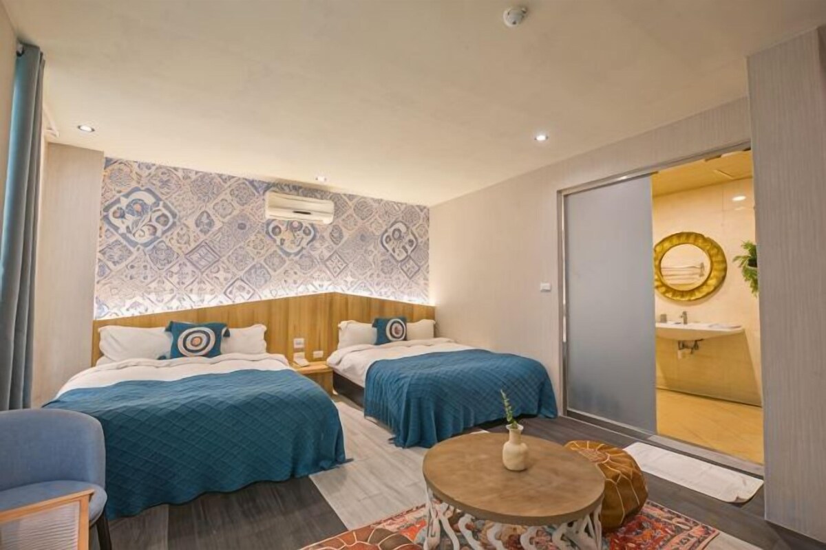 A hotel room with two beds, a small round table, a sofa, and a bathroom visible through a sliding door.