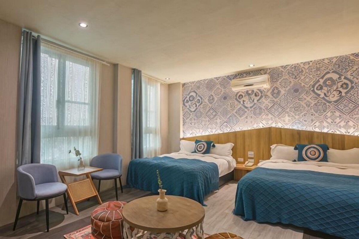 A hotel room with two beds, a small table, a chair, and a decorative wall.