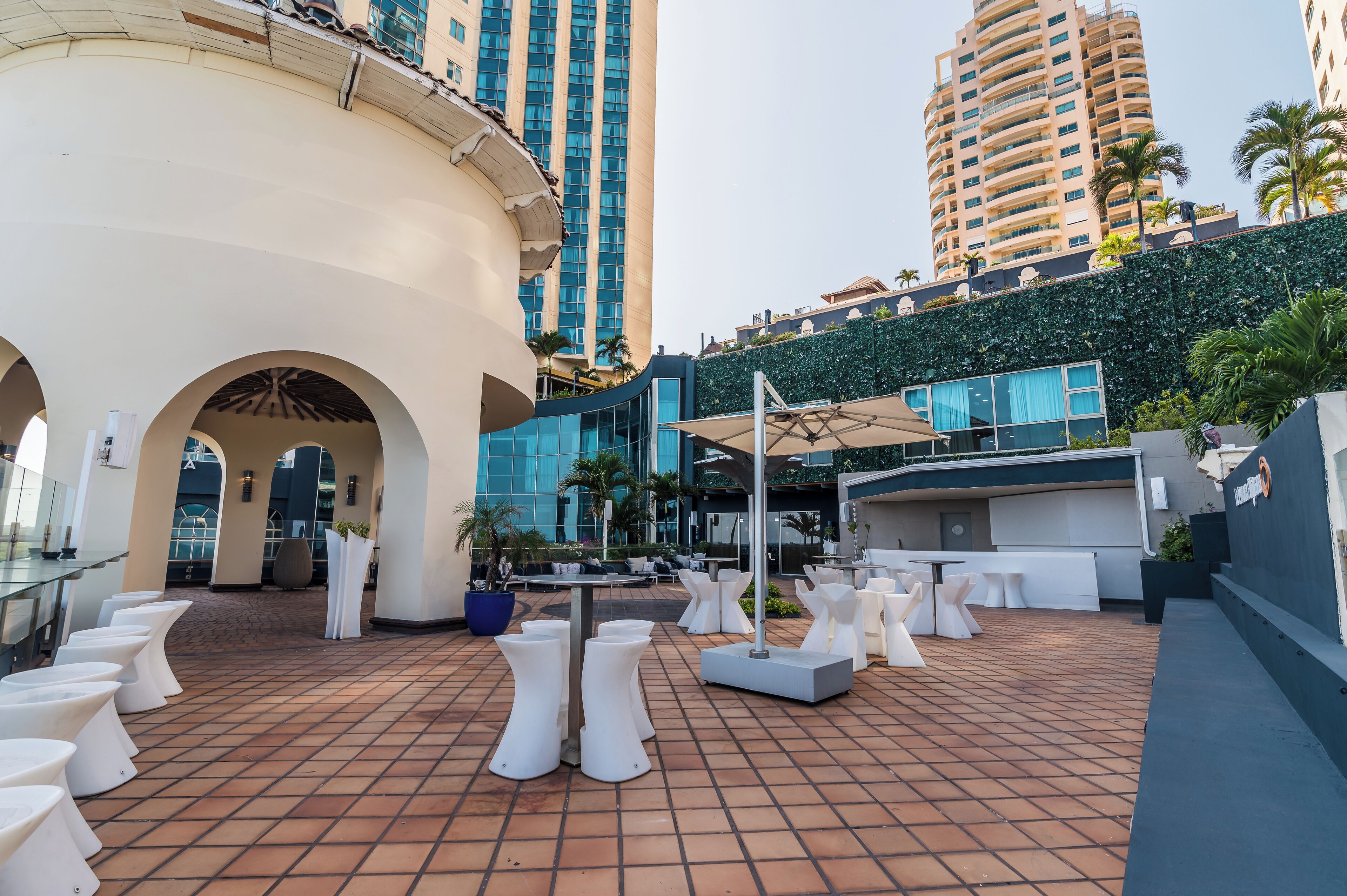 A rooftop terrace with white tables and chairs, a potted plant, and a view of tall buildings.