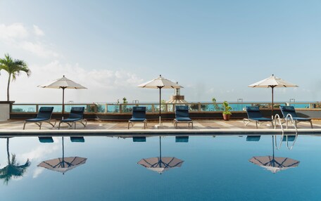 A poolside area with lounge chairs and umbrellas, a clear sky, and a view of the ocean.
