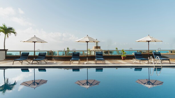 A poolside area with lounge chairs and umbrellas, a clear sky, and a view of the ocean.