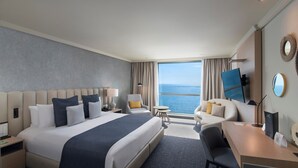 A modern hotel room with a large bed, a desk, a chair, a small table, and a view of the ocean.