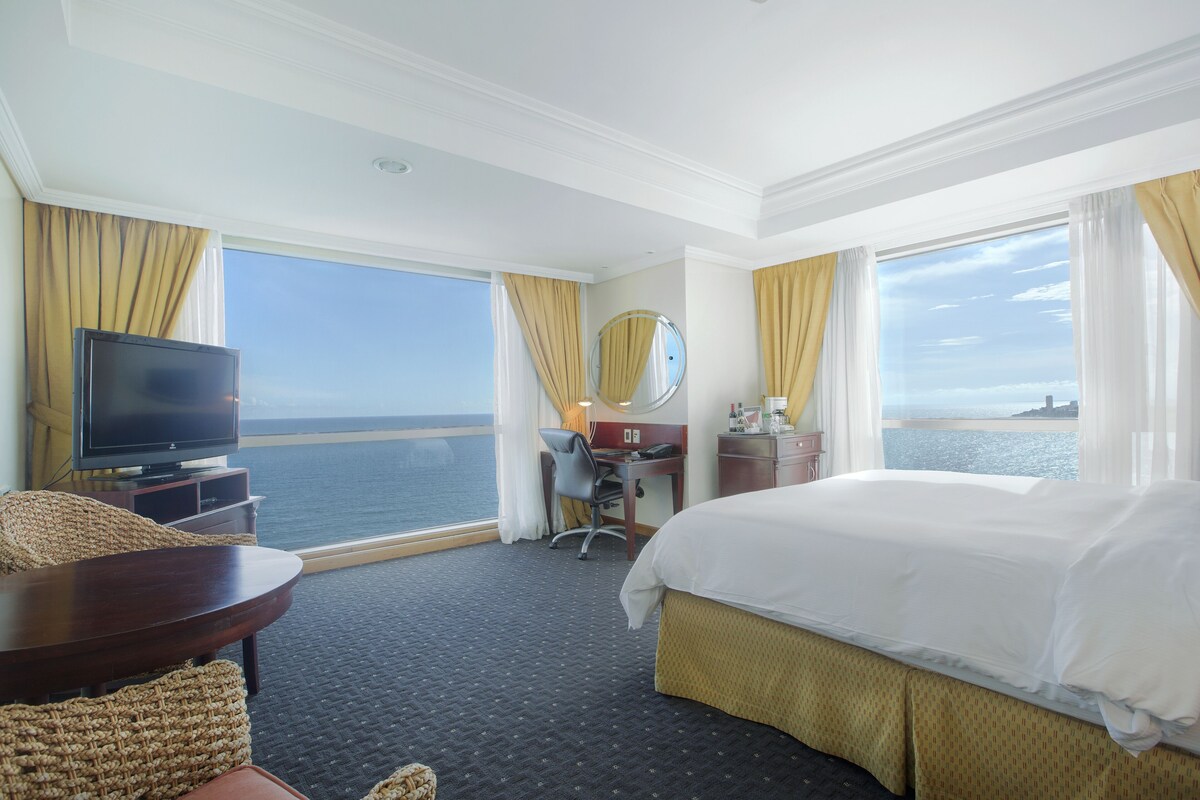 A hotel room with a bed, a desk, a chair, a television, and a view of the ocean.