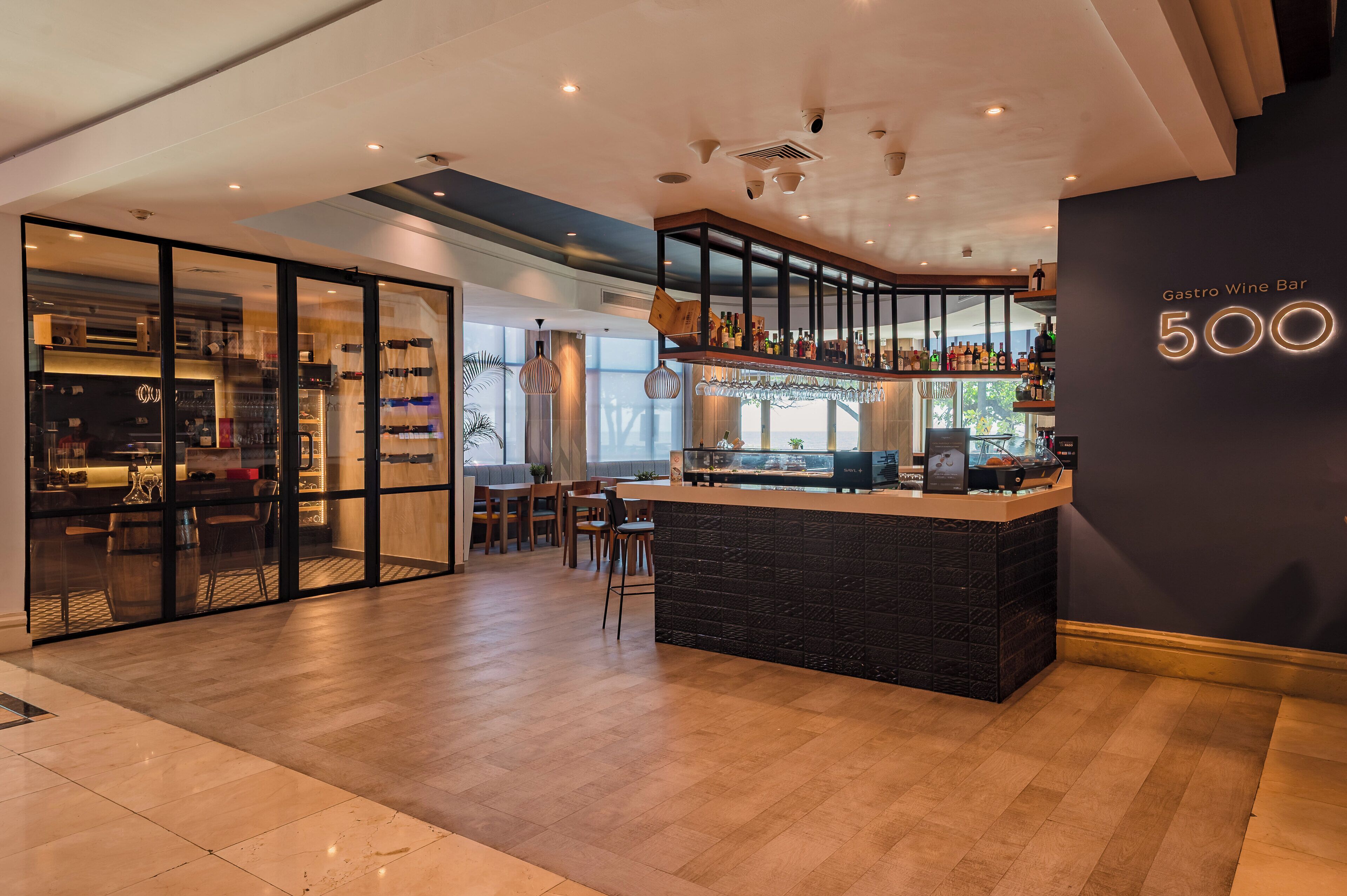 A modern bar area with a counter, seating, and a view of the adjacent restaurant.