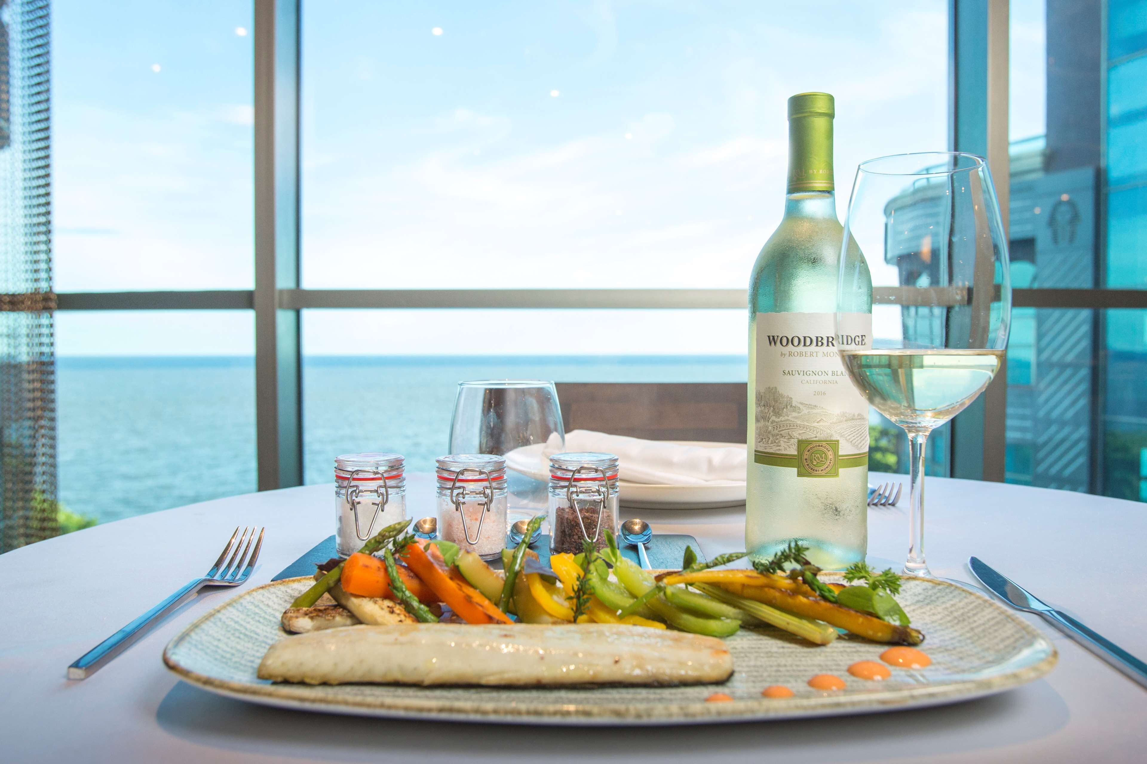 Una mesa con un plato de verduras a la parrilla y pescado, una botella de vino y una copa de vino, con vista al mar de fondo.