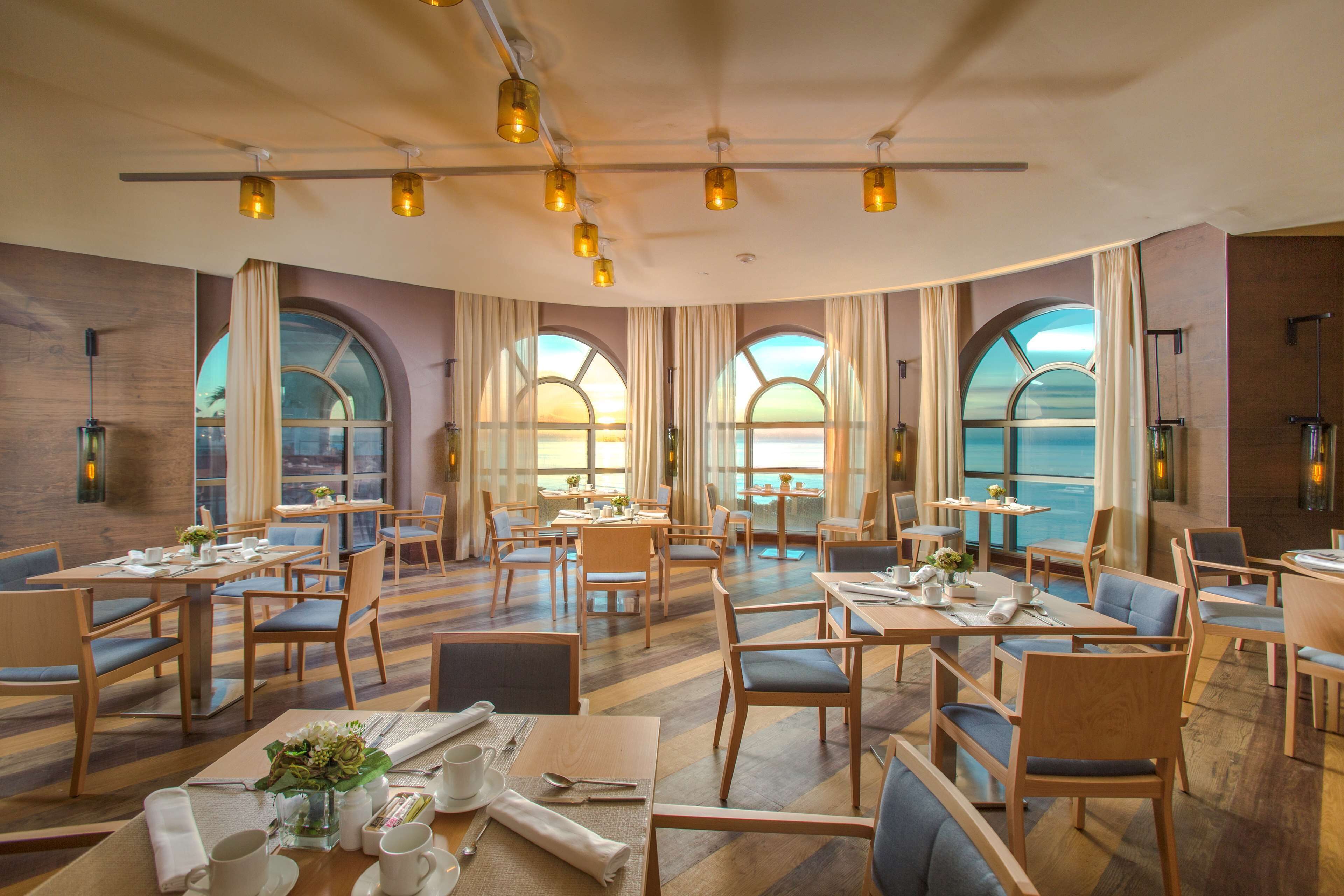 A spacious dining area with multiple tables and chairs, large windows with arched tops, and a view of the sea.
