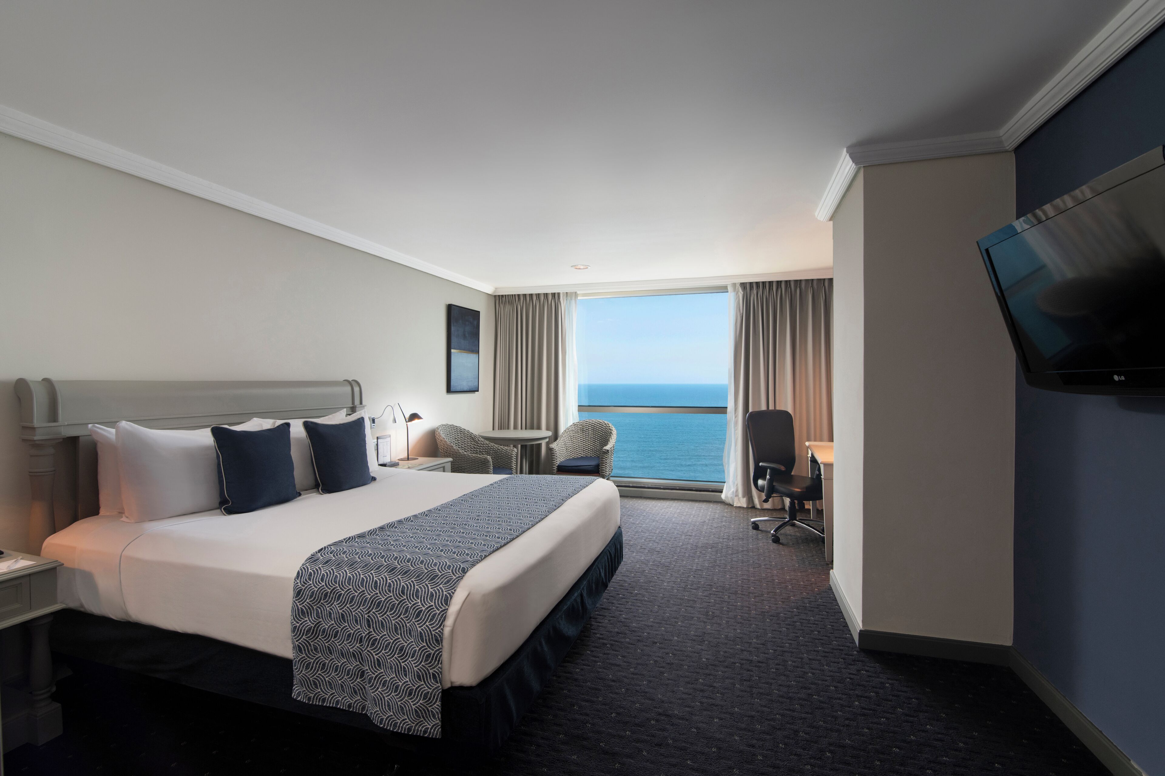 A hotel room with a large bed, a desk, a chair, a TV, and a view of the sea.