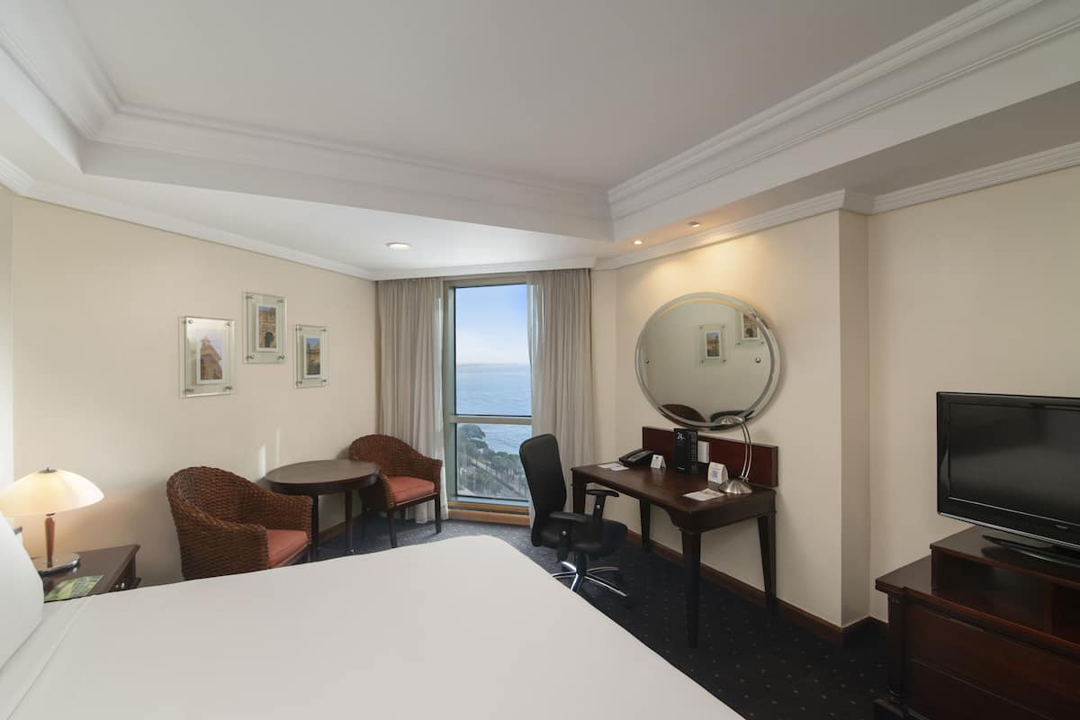 Une chambre d’hôtel avec un grand lit, un bureau avec une chaise, une télévision, une lampe de bureau, une chaise, une fenêtre donnant sur l’océan et des tableaux encadrés au mur.