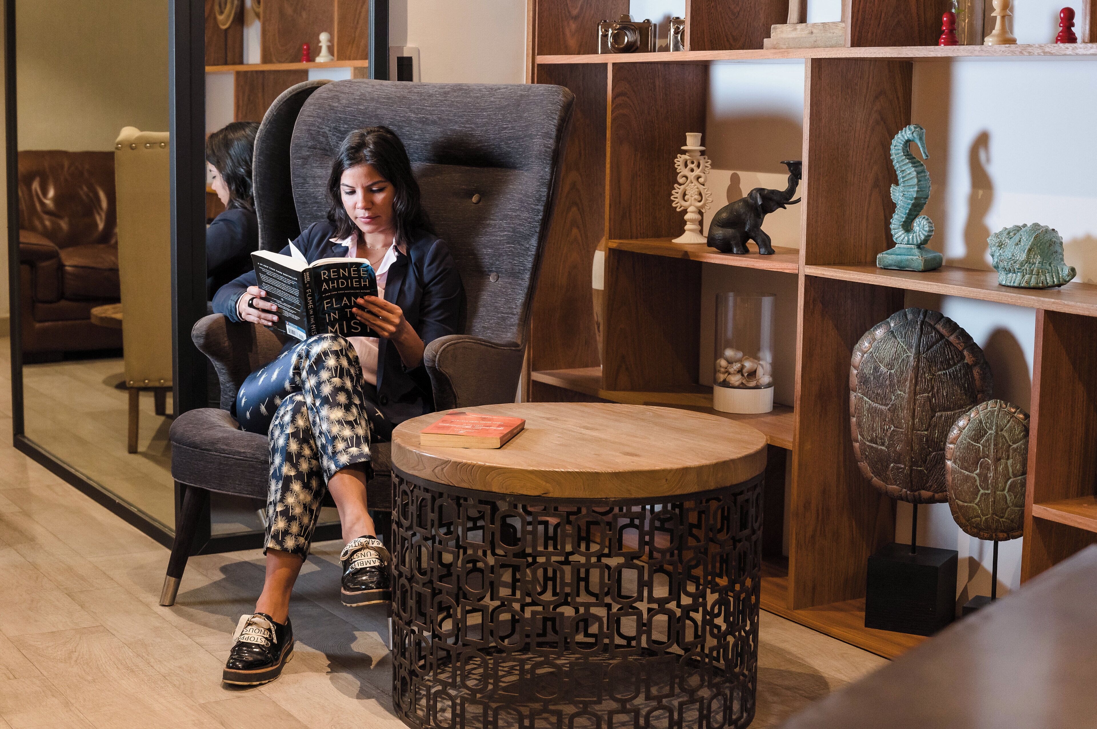 Una mujer leyendo un libro en un moderno espacio lounge con una mesa redonda de madera, sofá de cuero y objetos decorativos en estanterías.