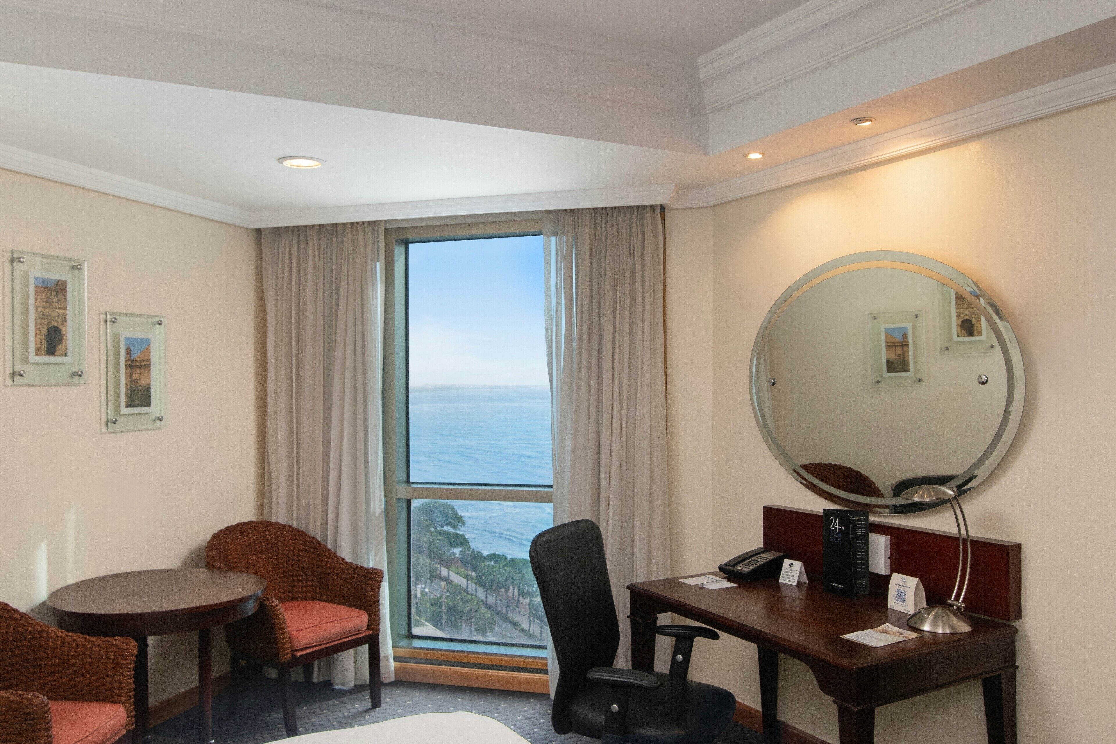 A hotel room with a large window, a round mirror, a desk with a telephone, and two chairs.