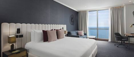A modern hotel room with a large bed, a desk, a chair, and a view of the ocean.