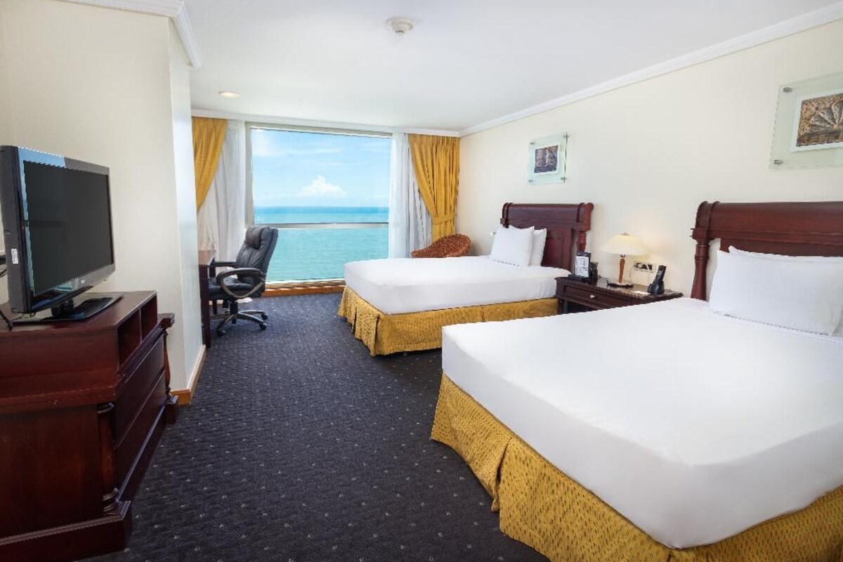 A hotel room with two beds, a desk with a TV, a chair, a window with a view of the sea, and a nightstand with a lamp.