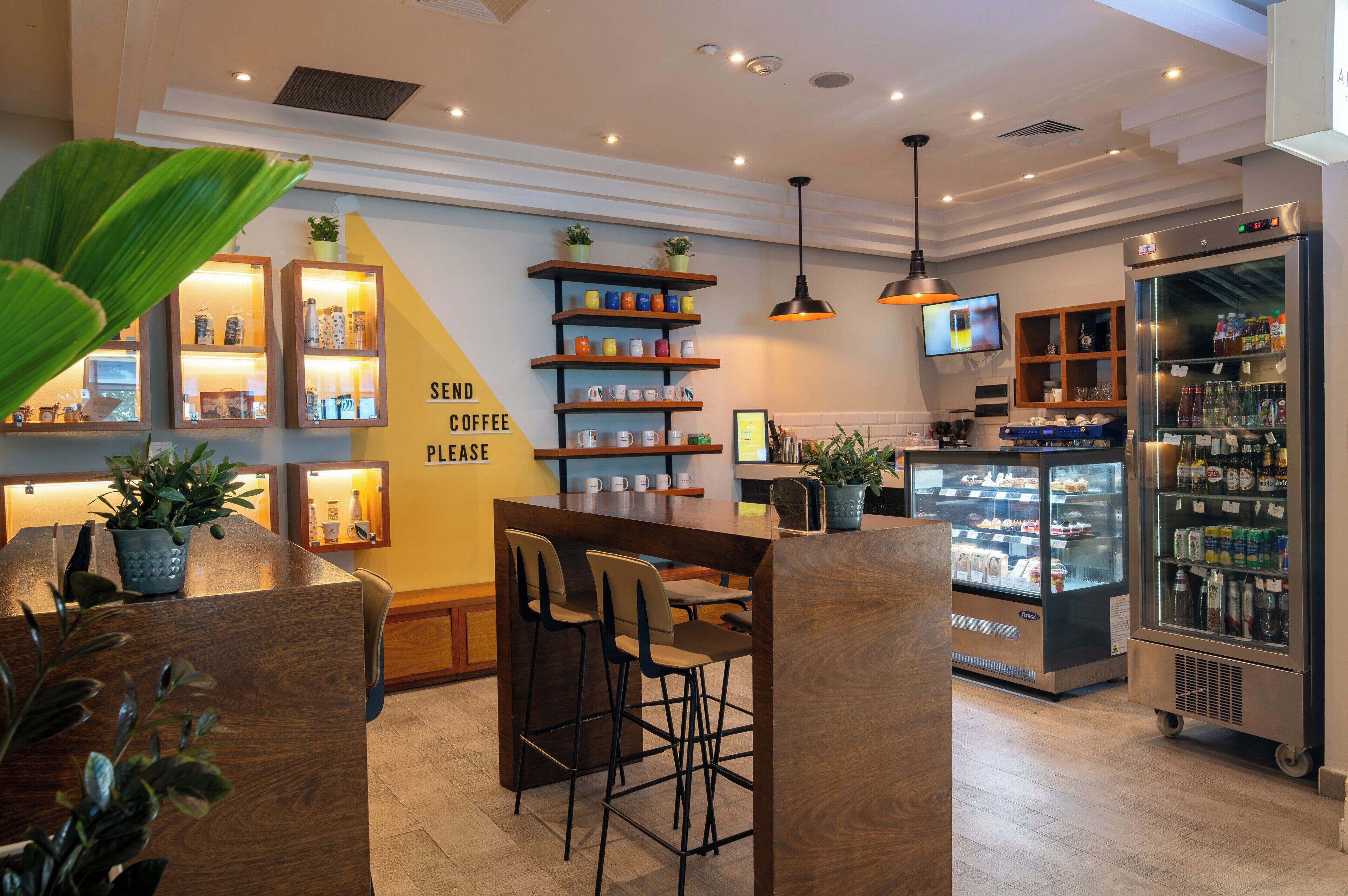 A modern coffee shop interior with a bar area, seating, shelves with coffee mugs, a refrigerator, and a television.