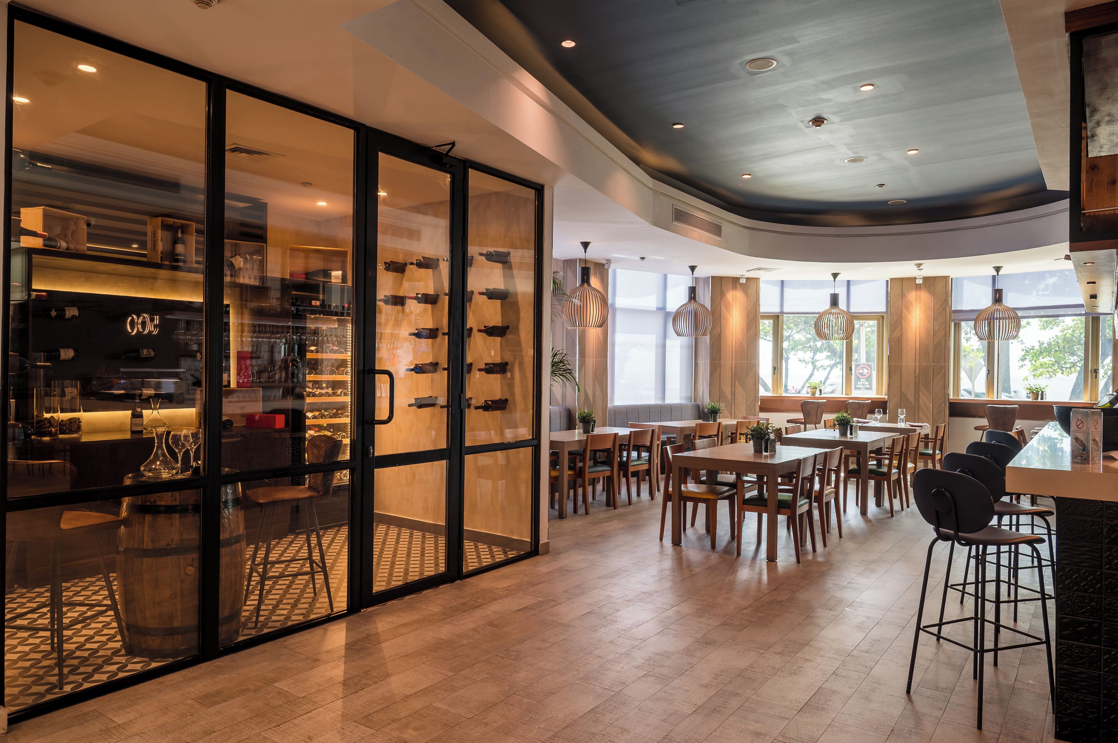 A modern restaurant with a glass partition separating the dining area from the kitchen. There are multiple tables and chairs arranged for dining, and the ceiling features recessed lighting.