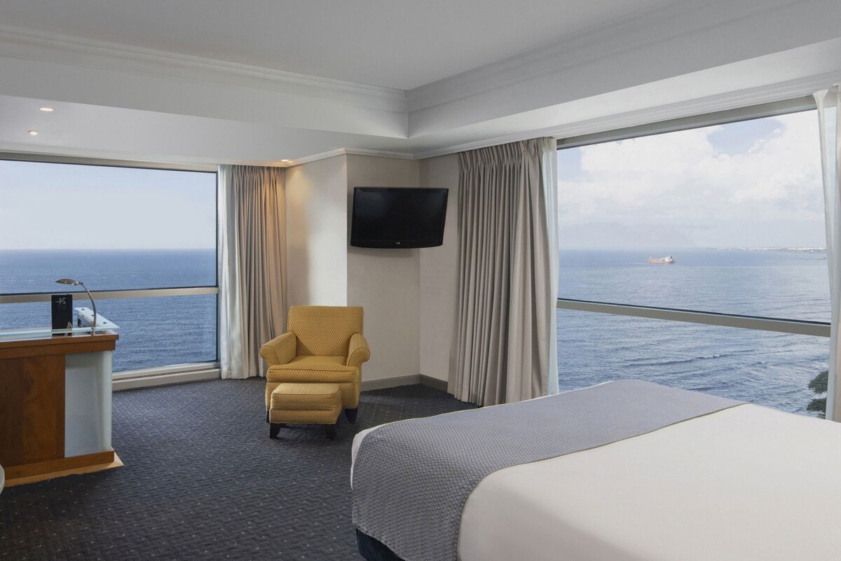 Une chambre d’hôtel équipée d’un lit, d’un bureau, d’une chaise, d’une télévision et d’une grande fenêtre donnant sur la mer.