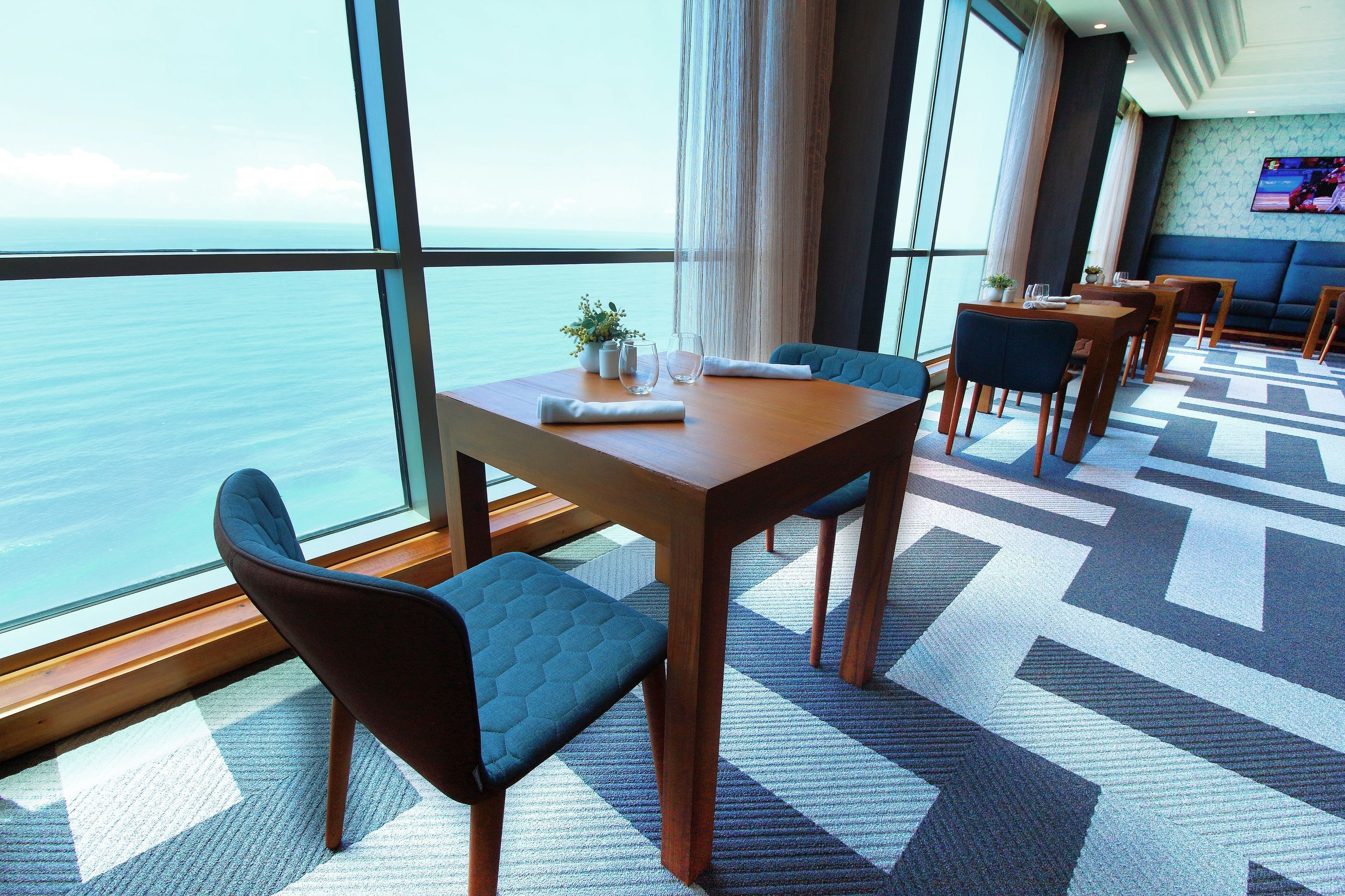 Un espace repas moderne avec une grande fenêtre donnant sur la mer, une moquette à motifs et un téléviseur fixé au mur.
