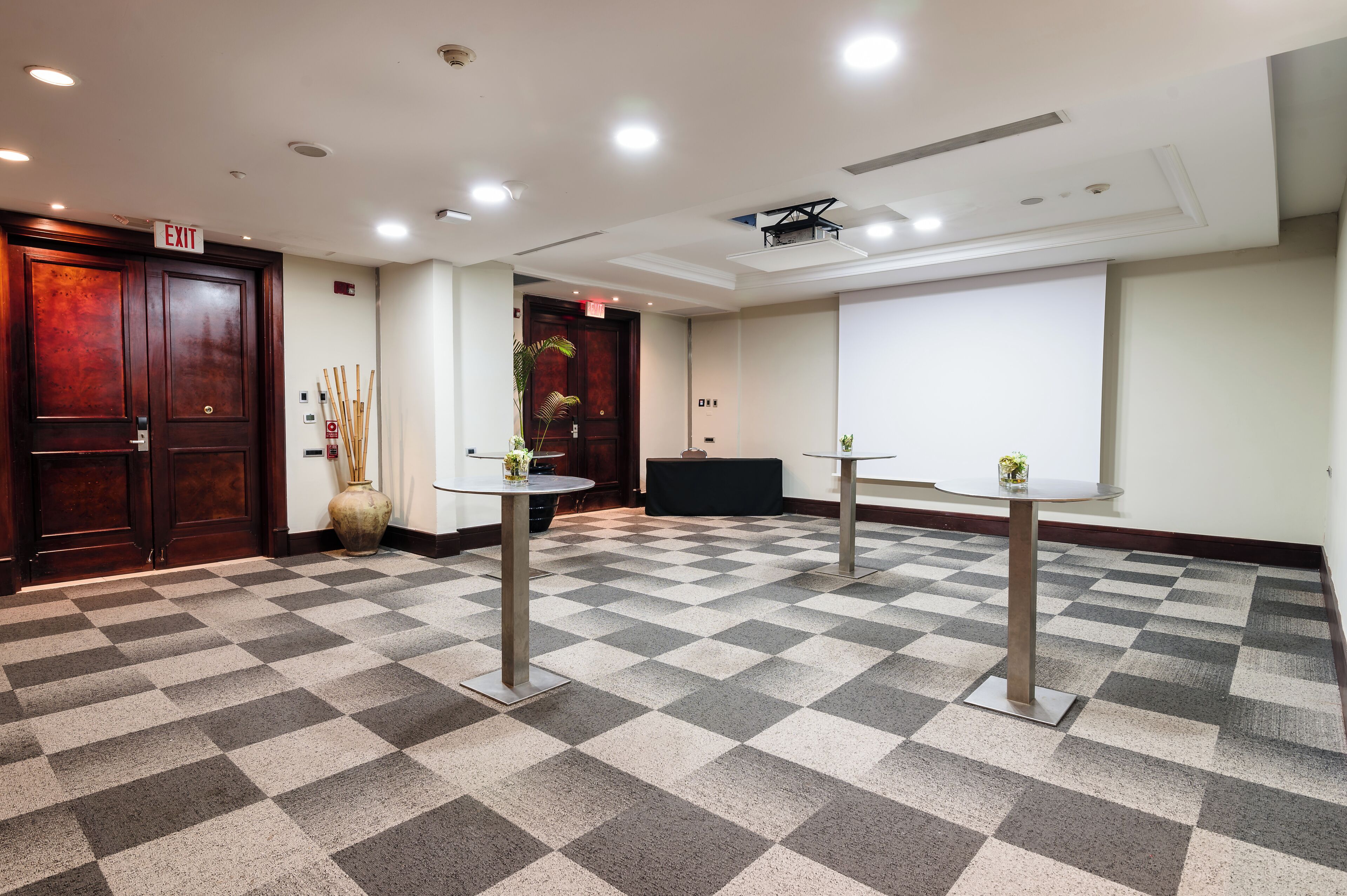 Une salle de conférence avec un sol à carreaux, deux tables ornées de plantes, un écran de projection et une porte de sortie.