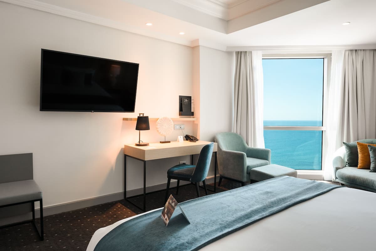 Une chambre d’hôtel comprenant un lit, un bureau avec une chaise, une télévision et une vue sur la mer grâce à une grande fenêtre.