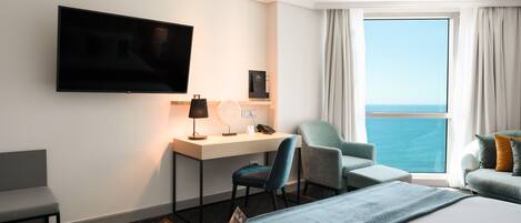 A hotel room with a bed, a desk with a chair, a TV, and a view of the sea through a large window.