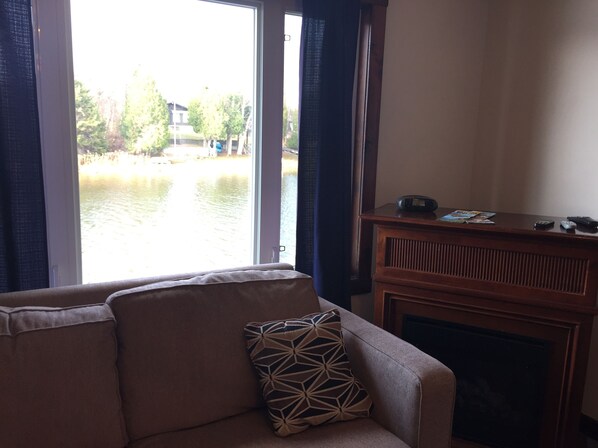 Un salon avec un canapé, un coussin à motifs, une cheminée et une vue sur une étendue d’eau, visible à travers de grandes fenêtres.