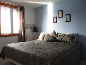 Un dormitorio con cama, ventana con cortinas, lámpara y tres cuadros en la pared.