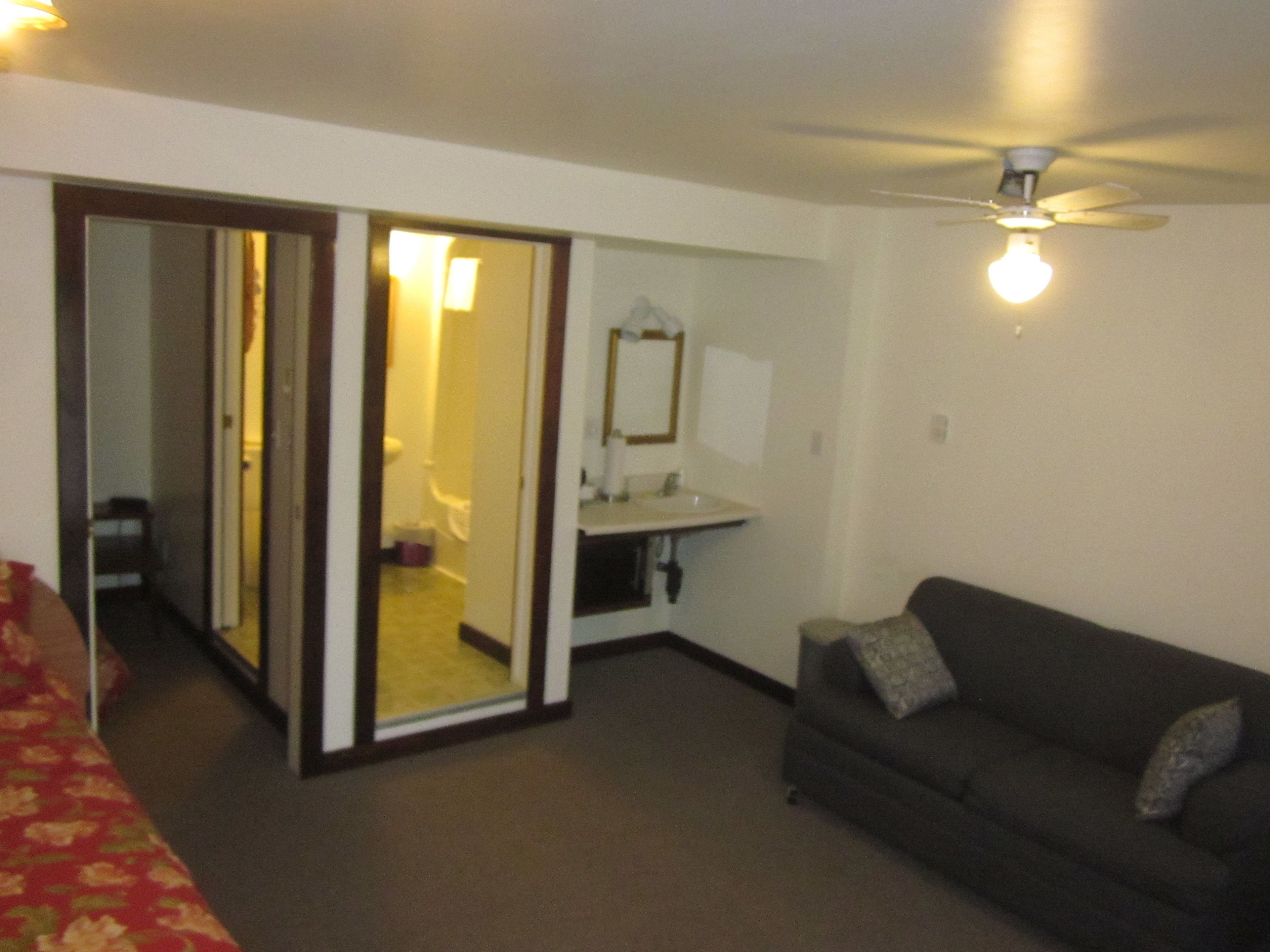 Habitación de hotel con cama, sofá, baño con espejo y ventilador de techo.